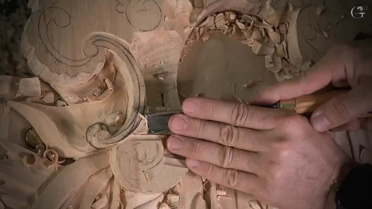 Both hands press a gouge toward the pencil line in the lowered wood near the scroll
