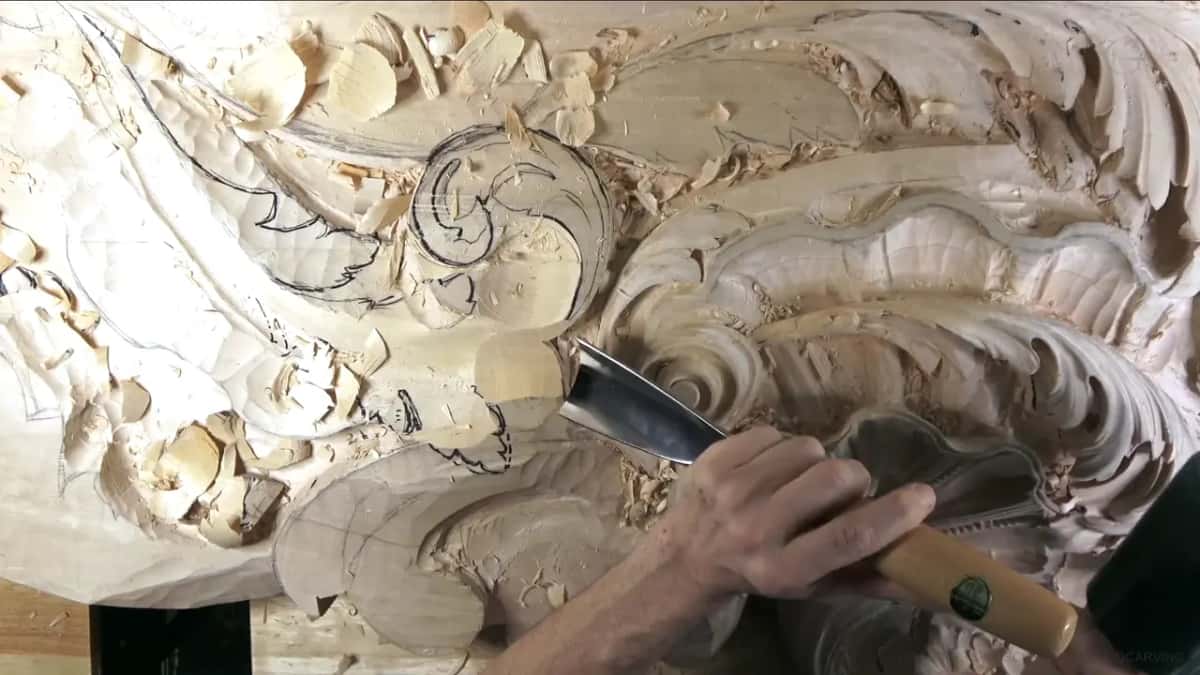 Both hands on a big gouge beside the scroll, chips piled all around it.