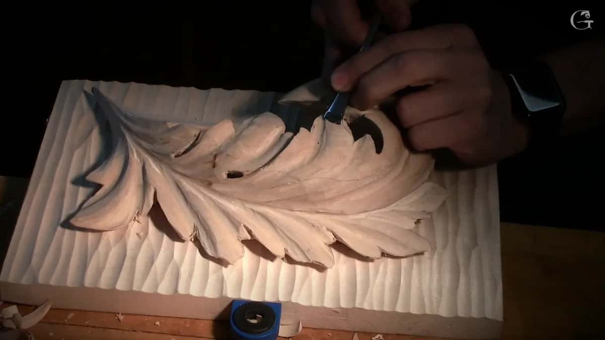 In low light, a hand works a gouge near the curl while the shadows fall to the left