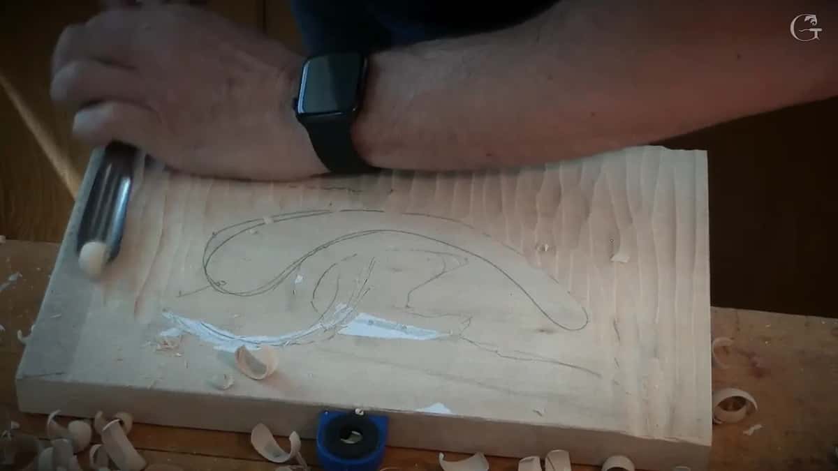 A hand holds a gouge across the board, rows of tool marks already running down the right side