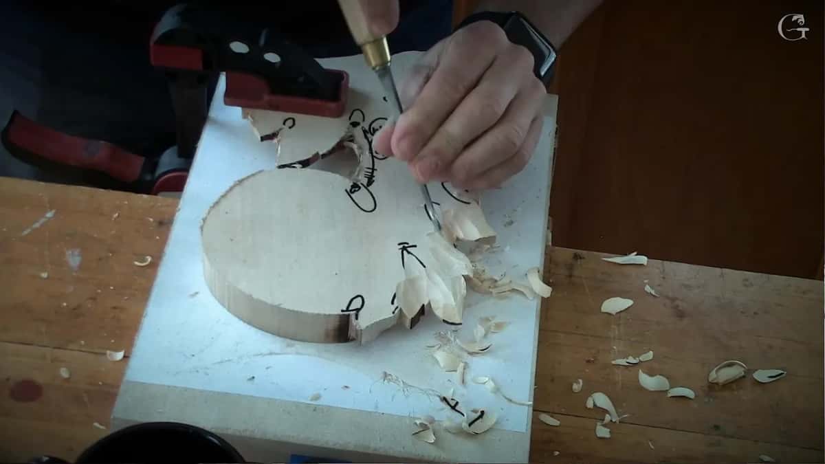 Thin gouge at a leaf tip, one hand guiding the blade from above, shavings on the board