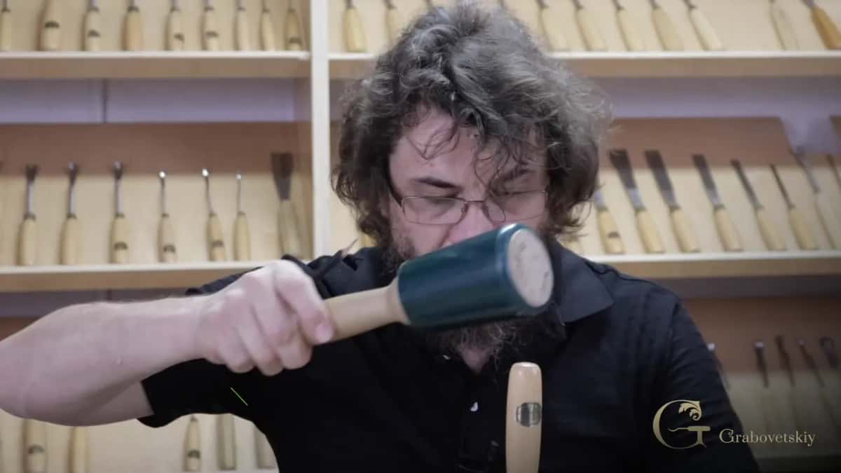 Mallet swung hard at a gouge, with rows of carving tools racked on the wall behind me.