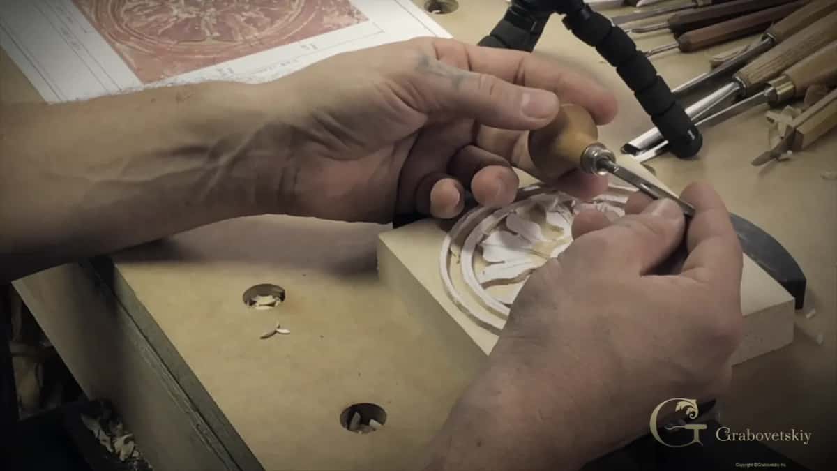 Both hands on a round-handled palm tool, with the printed reference picture and a rack of gouges behind.
