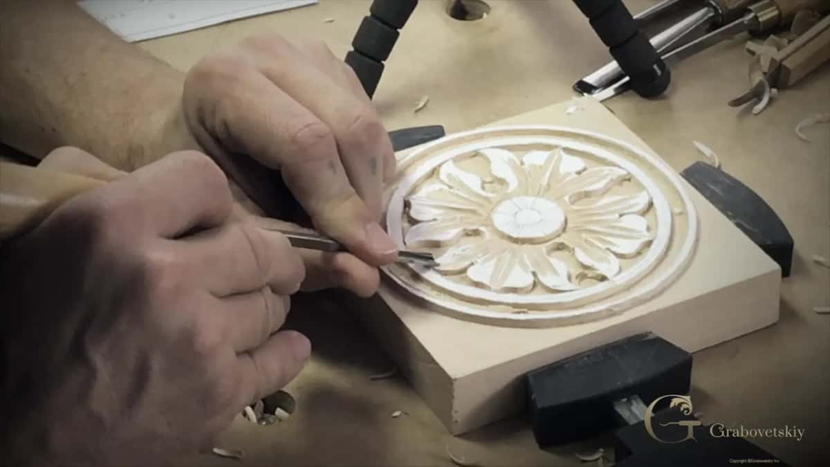 Seen from the side, two hands guide a gouge at the rim of the rosette while more gouges lie on the bench behind.
