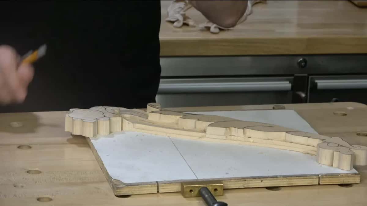 Pencil raised over the long carving, which lies on a white board held by a brass clamp.