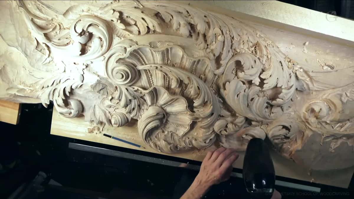 One hand steadies the long carved panel while a black hair dryer aims at its lower edge
