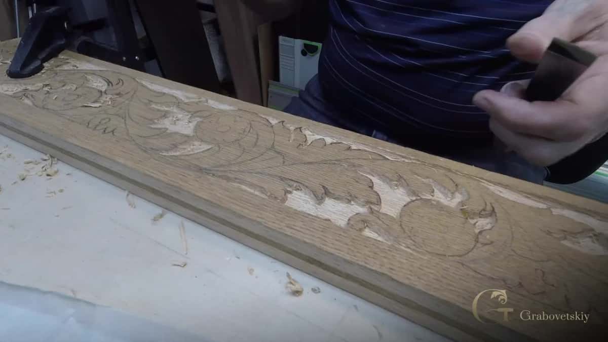 A long carved board with leaves lies across the bench while a hand at right holds a dark tool.