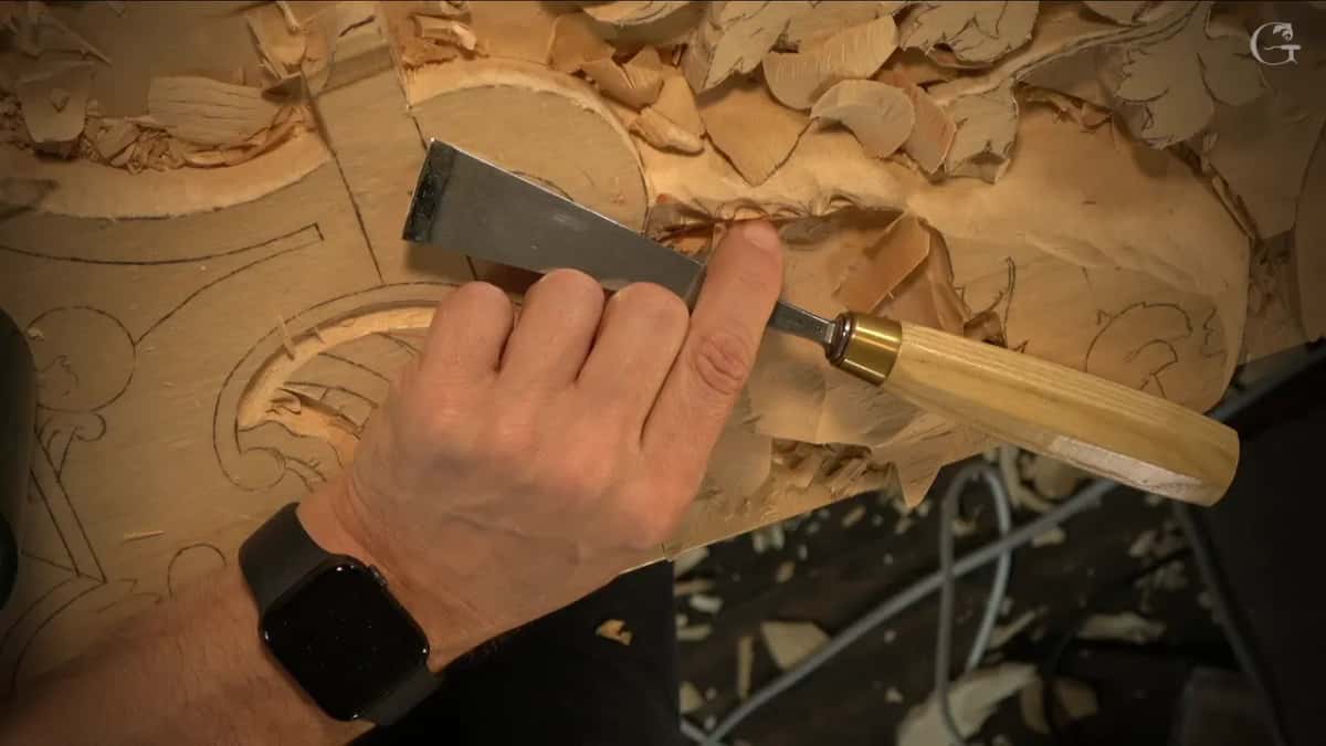A right hand grips a flat chisel with a brass ferrule above the carved curves and leaves.