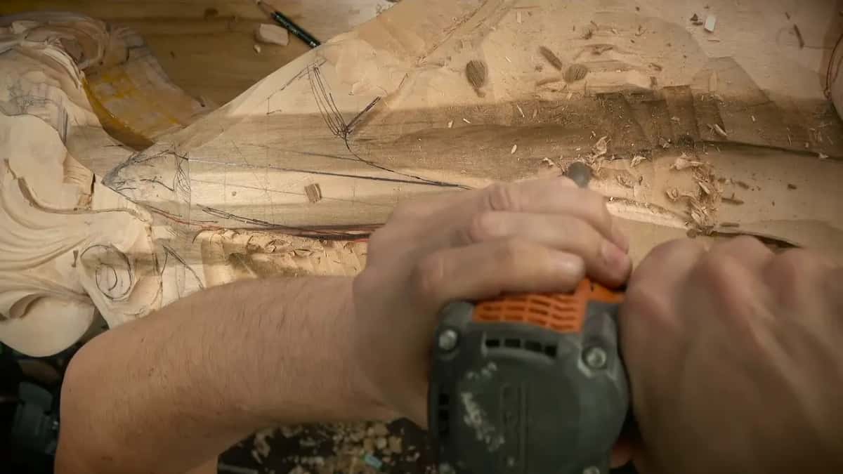 Two hands hold an orange and gray drill pointed down into the wood beside a drawn scroll