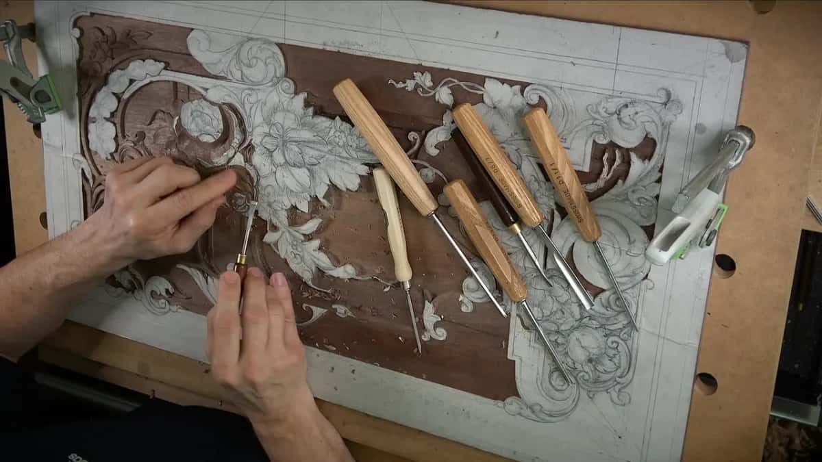 Panel from above with gouges laid across it, one hand steadying the board while the other works a small tool