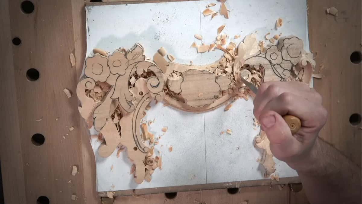 From above, a hand pushes a wood-handled gouge into the panel