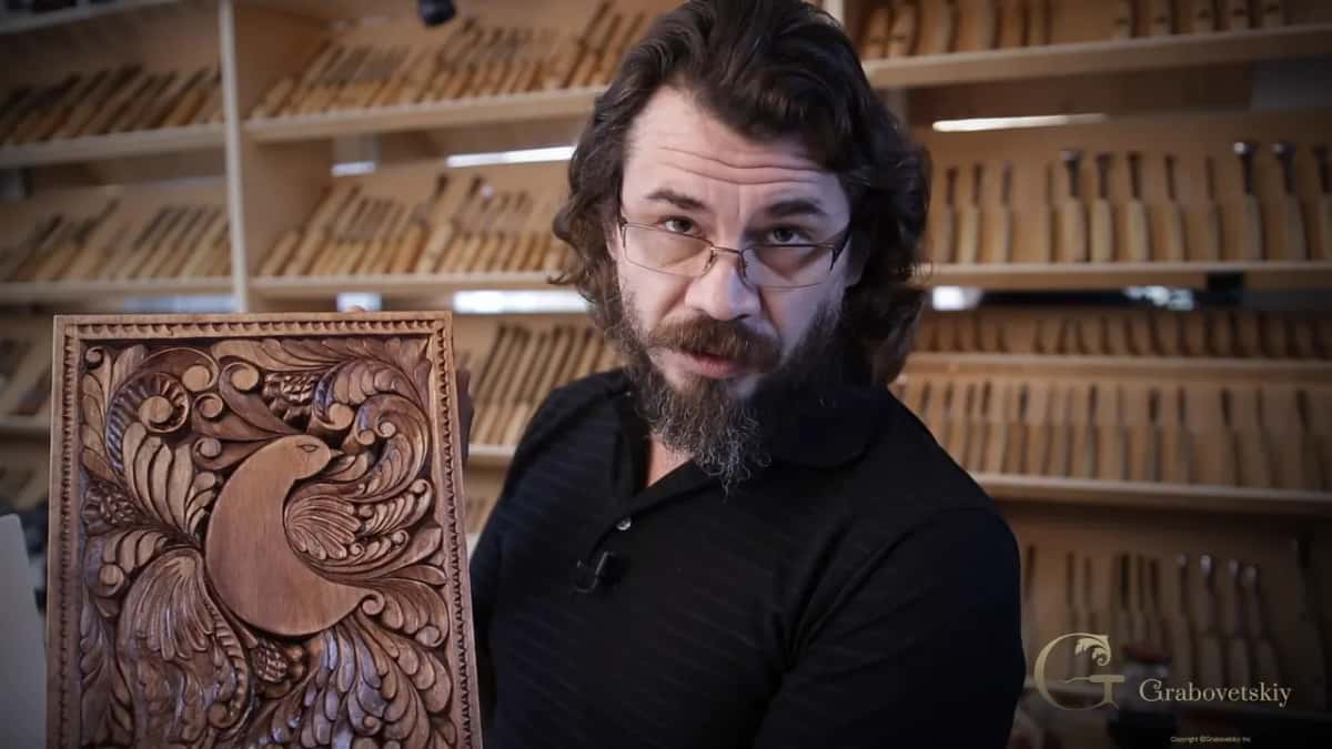 Alexander holding up the finished board, carved, stained with walnut shells and oiled, in front of his tool rack.