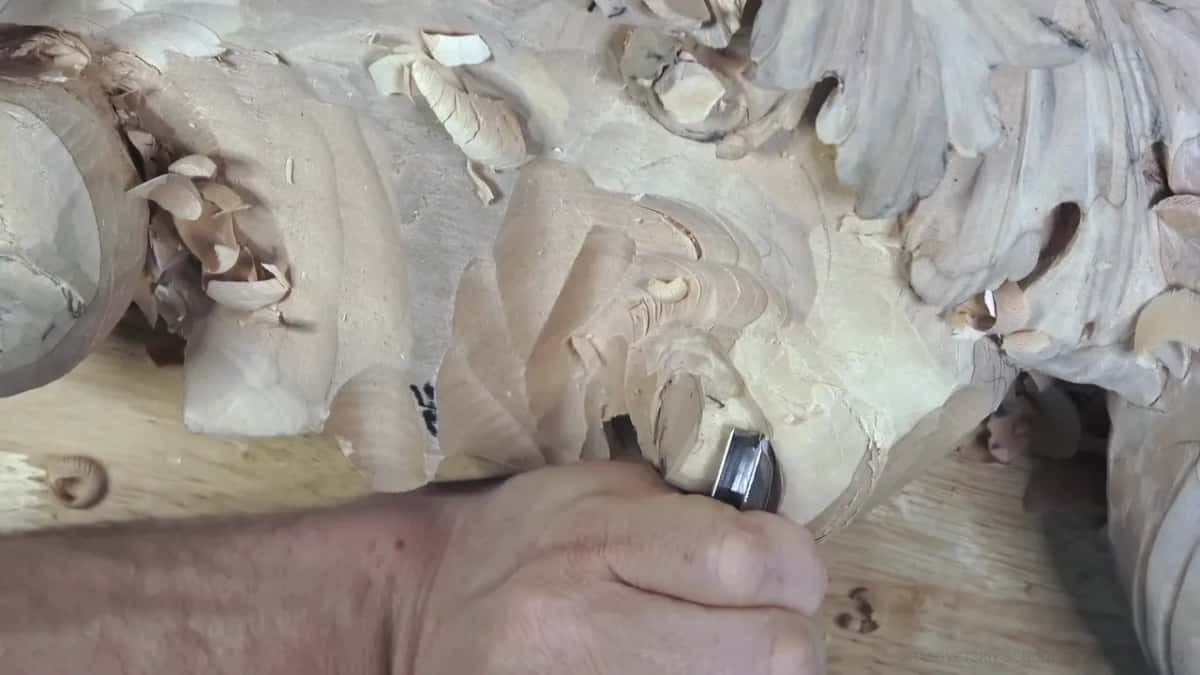 One hand drives a gouge around the rolled body of the volute, and the leaves above it are already carved.
