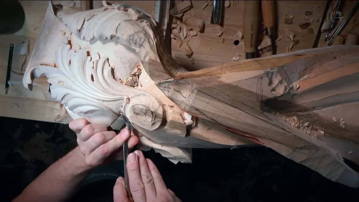 Under brighter light, both hands steer a narrow tool into the curl of the volute, leaves at left