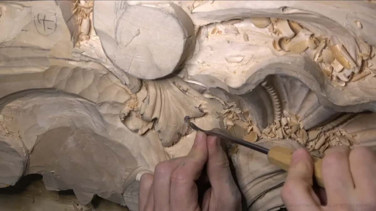 Both hands steer a yellow-handled gouge into the hollow beside the beaded shell, chips piled all around.