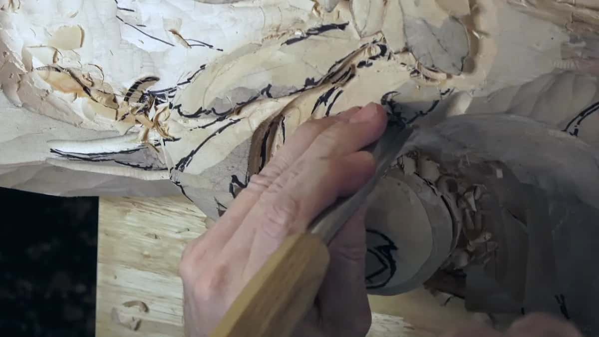 A right hand drives a wood-handled gouge into the rolled lobe, with chips caught in the curl.