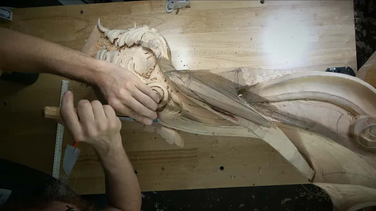 Both hands on a gouge where the acanthus meets the side, a ruler standing at the left edge