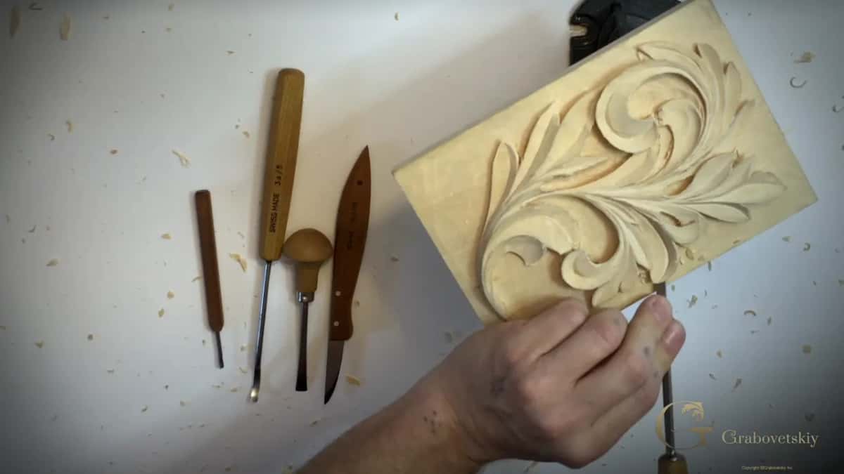 Four carving tools laid out in a row, and a hand steadying the acanthus panel just beside them