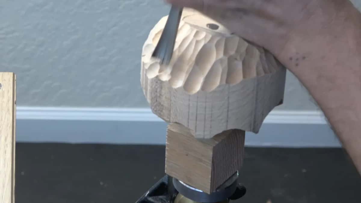 A hand drives a gouge down from the top edge of the mounted blank, rows of scallops below