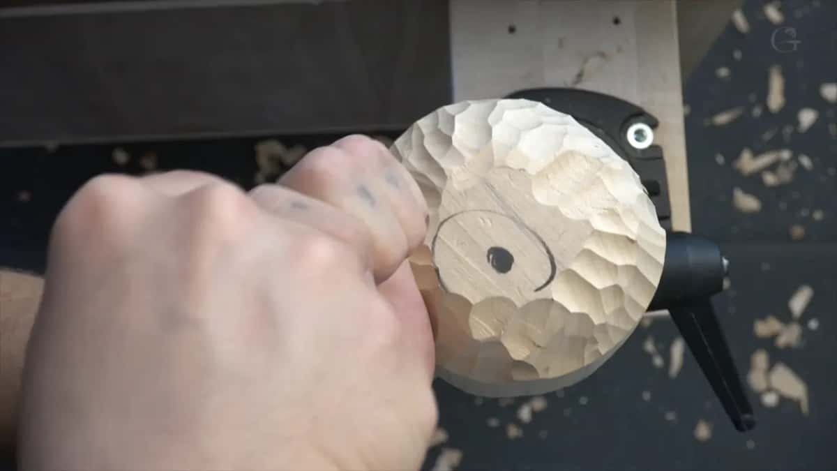 Seen from above, two fists drive a gouge into the scalloped ball, a circle and dot drawn on its top