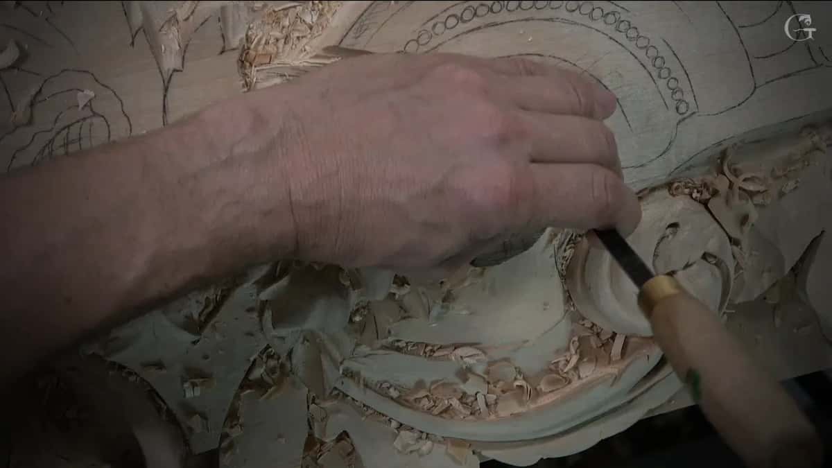 Right hand on a pale handled gouge, its blade down in the curl of the scroll among shavings