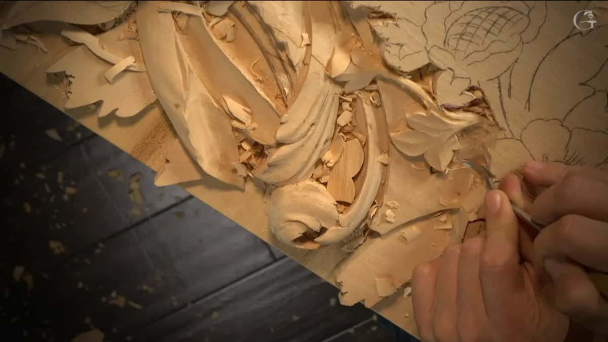 Both hands steady a fine tool at the carved leaves while the pencil flower waits.
