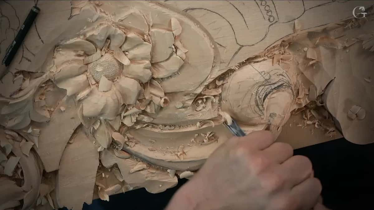 A hand holding a narrow gouge at the swirl beside the carved flower, with a pencil lying at the top left