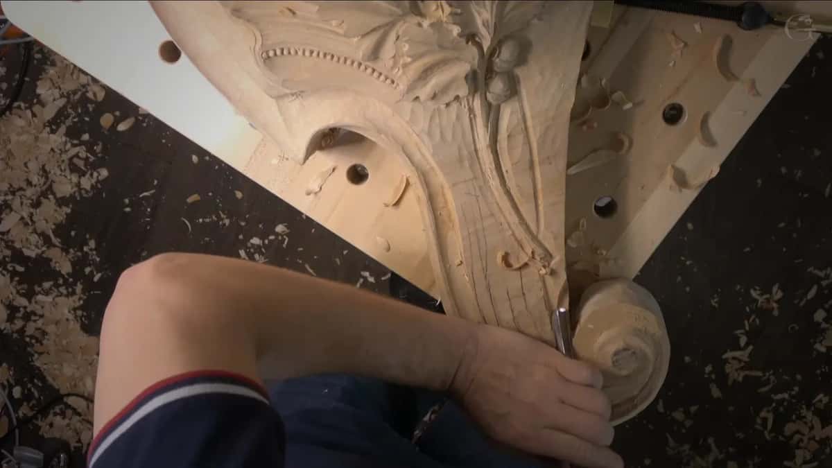 Seen from the other end, an arm holds a gouge low on the carving near the scroll's round end.