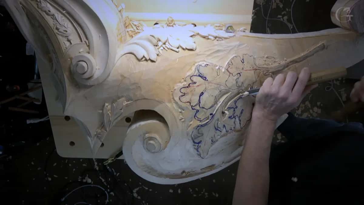 The whole scrolled piece seen from above while one hand drives a gouge into the leaves