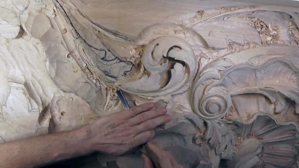Flat against the board, one hand presses a gouge along the outline beside the scroll.