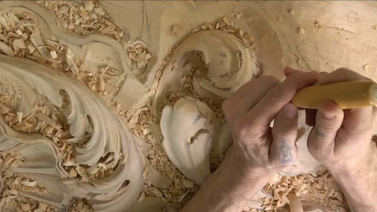 Knuckles bent around a pale handle, the gouge biting into the middle lobe while big scrolls sweep to the left.