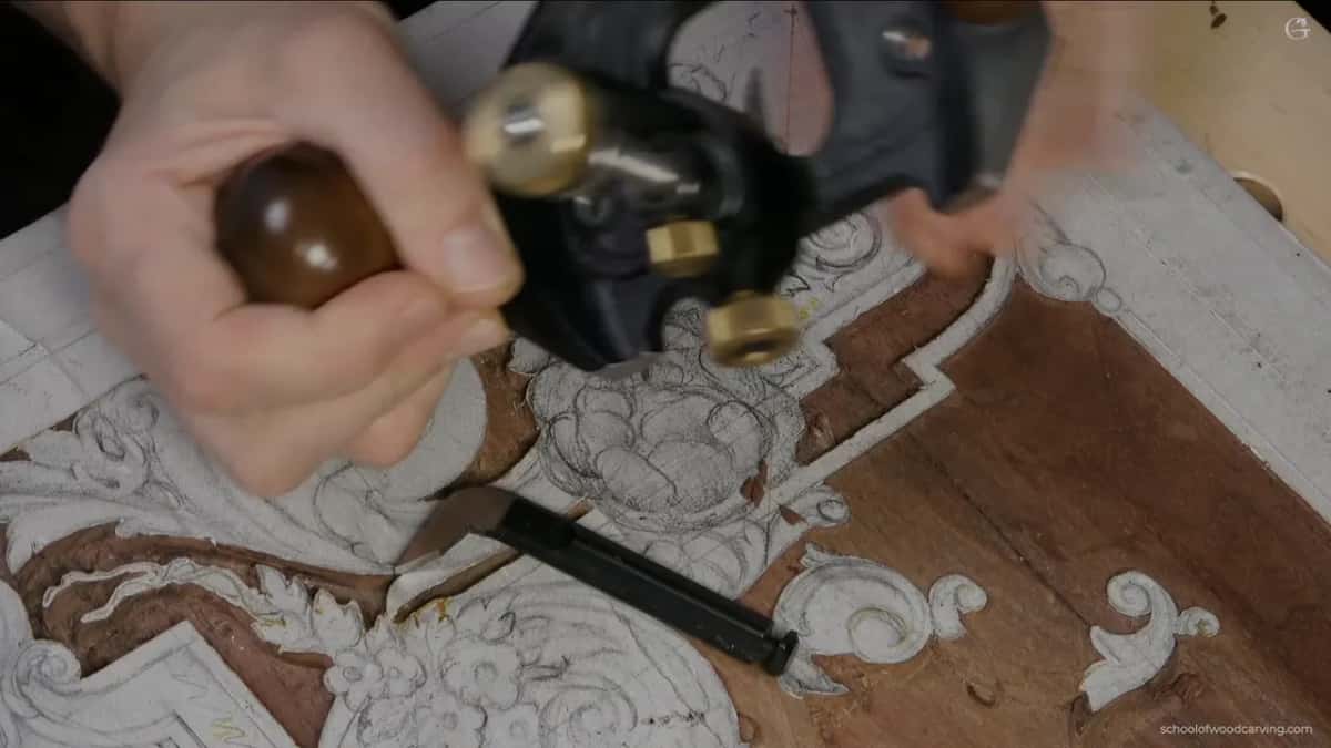 Close on the router plane, one hand on a wood knob and a gouge lying on the panel below