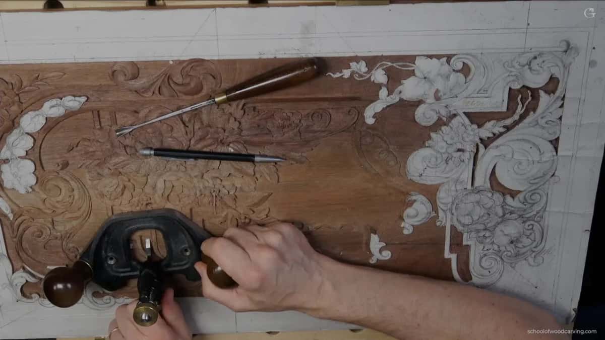 From above, a gouge and a pencil on the panel, both hands holding the plane at its bottom edge