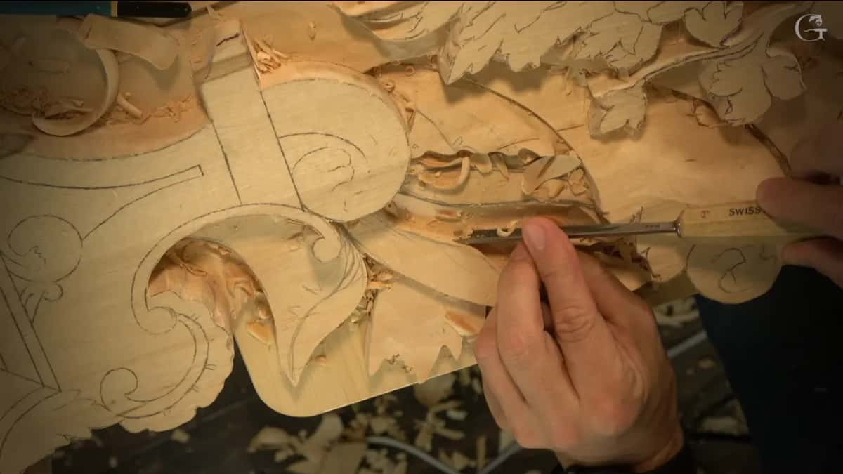 Fingers guide a narrow gouge deep inside a hollow, with pale chips piled around it and leaves above.