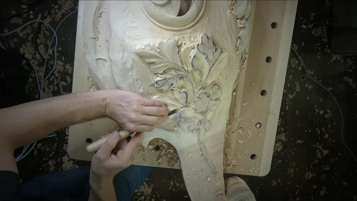 Two hands guide a small gouge across the leaves while the whole block sits on a bench top full of holes.