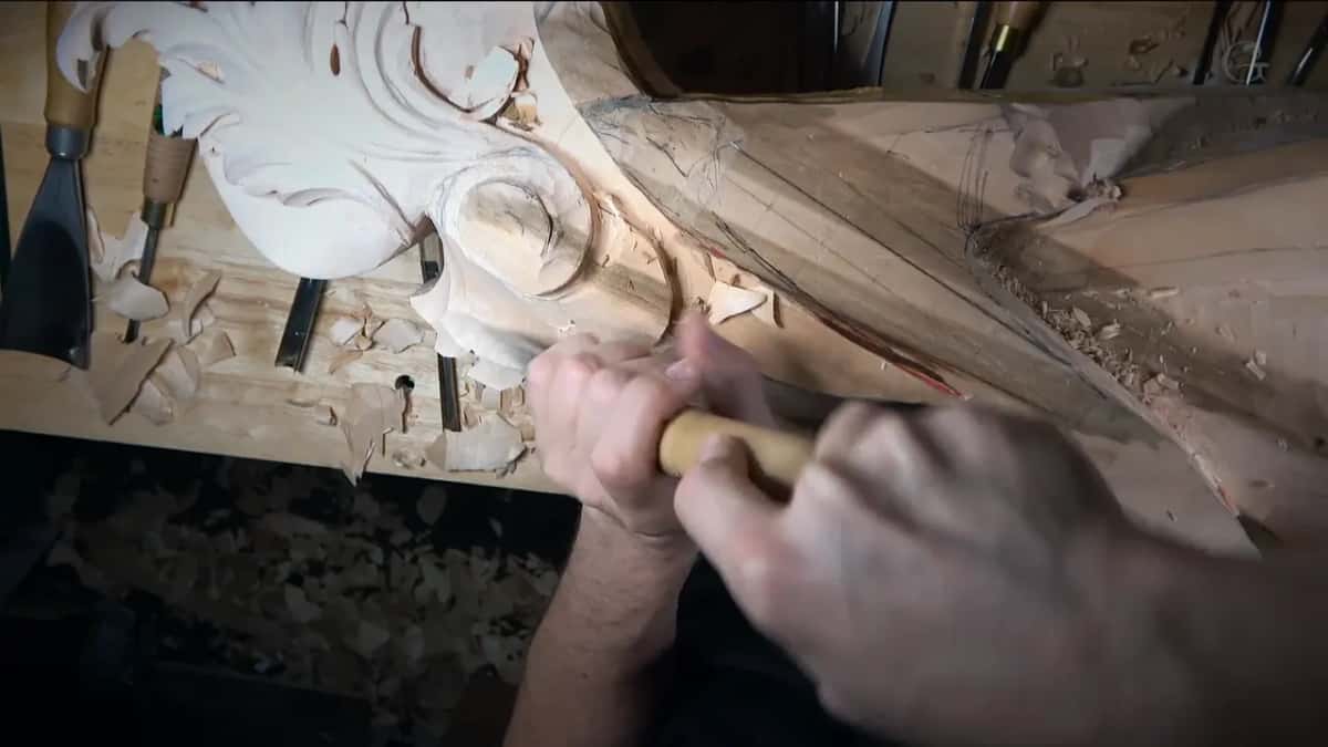 Two hands push a wide gouge with a pale handle across the slope, curls of shaving around it