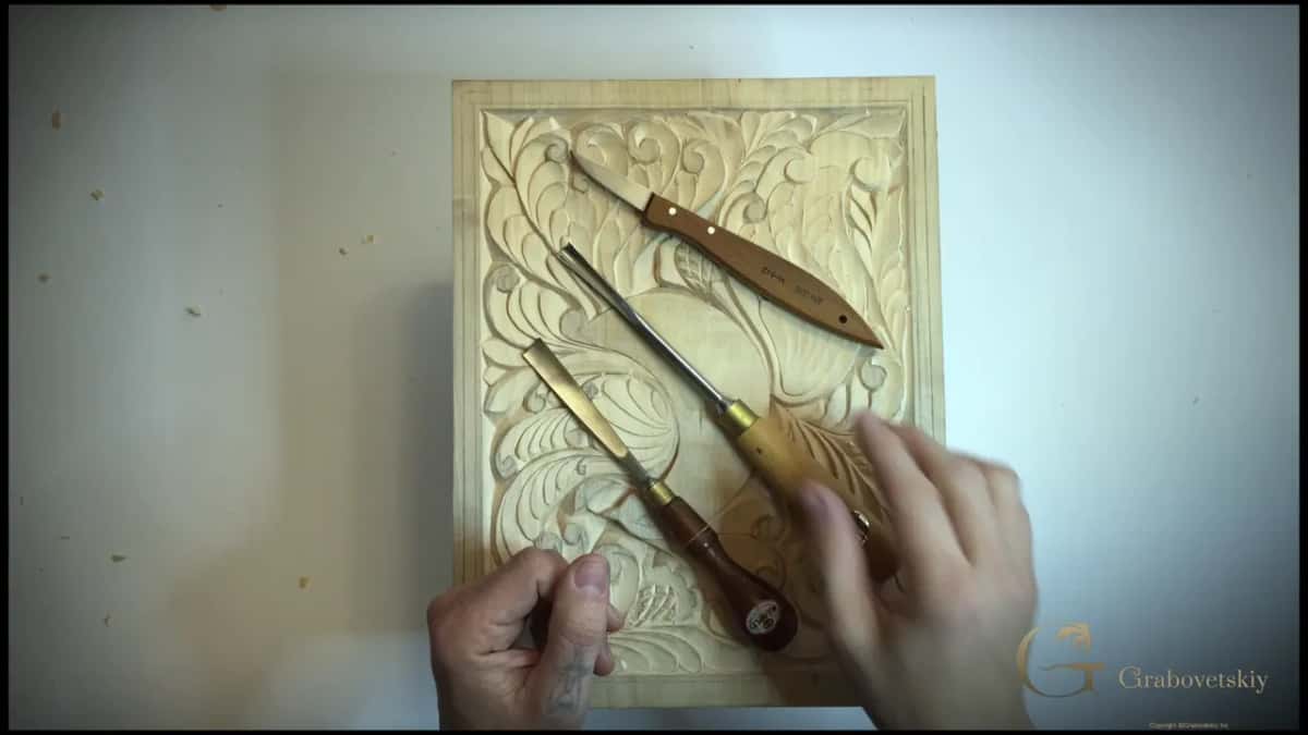 Three tools on the carving, the knife above, a dark-handled flat chisel and a pale-handled gouge below