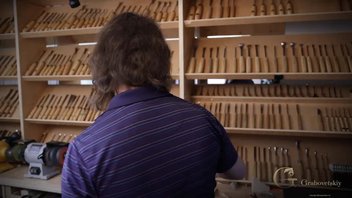 Seen from behind, reaching up to a wall of shelves full of carving tools, a bench grinder at the left