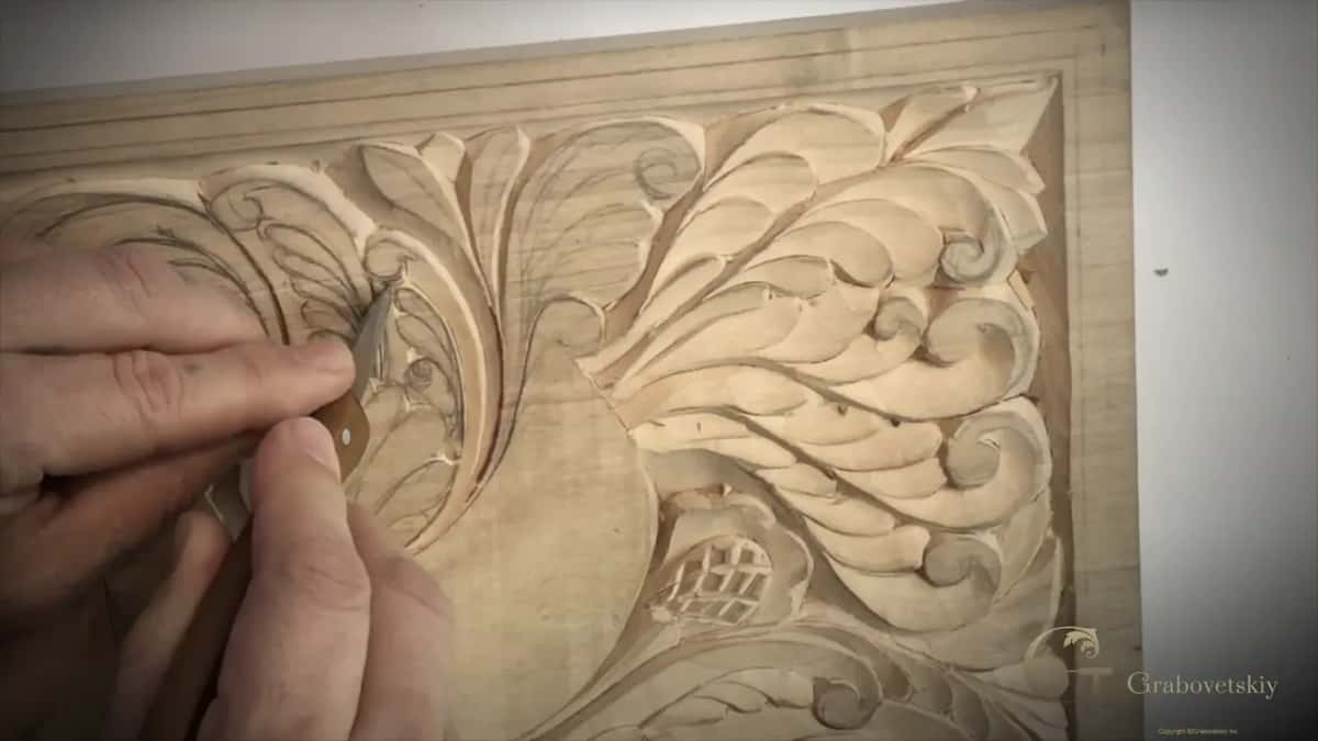 Fingers pinch a small knife at a carved curl on the turned board, the bird's round body beside it