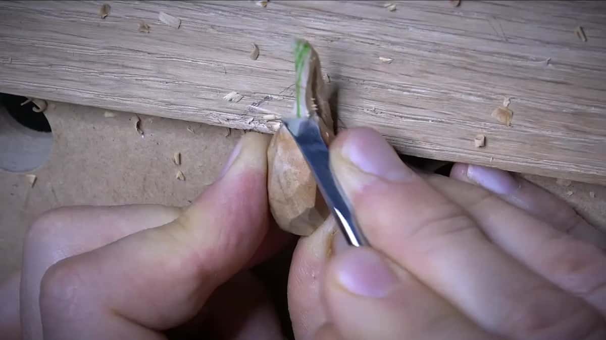 A knife shaves a tiny olive pressed against the edge of a board, a strip of green tape beside it