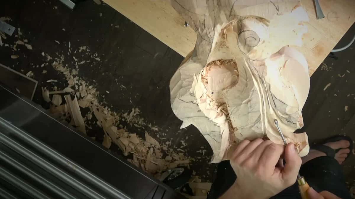 Thin tool held upright against the carving, right hand pinching the handle close to the wood
