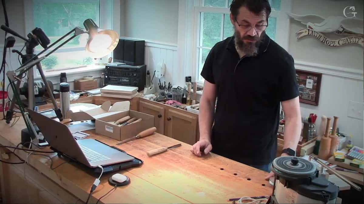 Standing at the bench with one palm flat on the top, a box of gouges at the far left