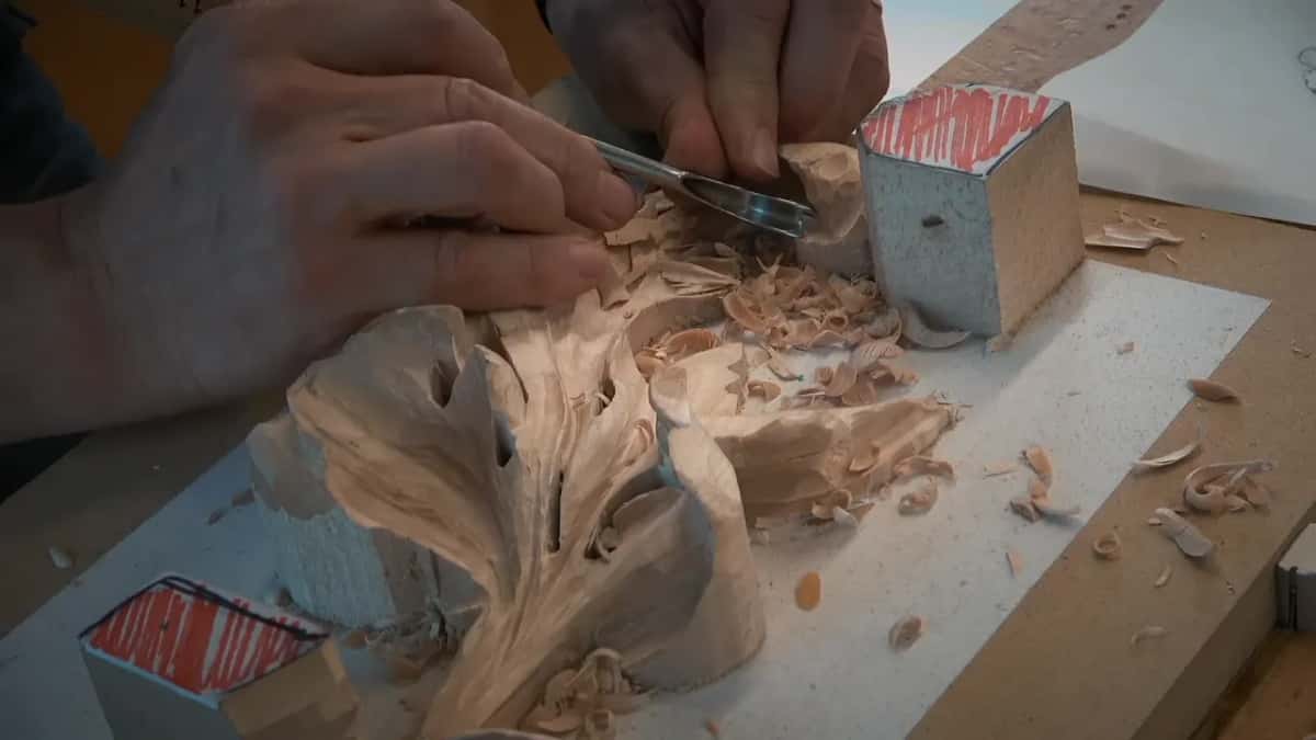 Close in, two hands work a tool into the top corner while curls of wood pile up
