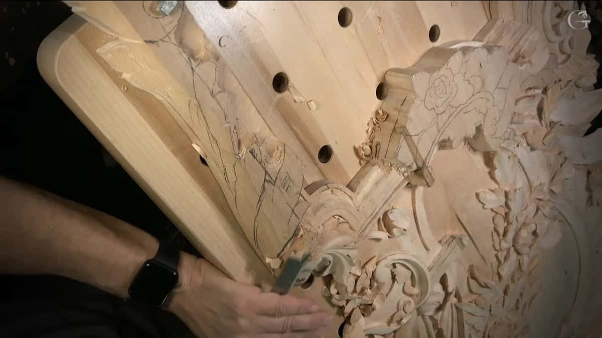 Seen from the side, a hand works a small tool into the carving below the flowers