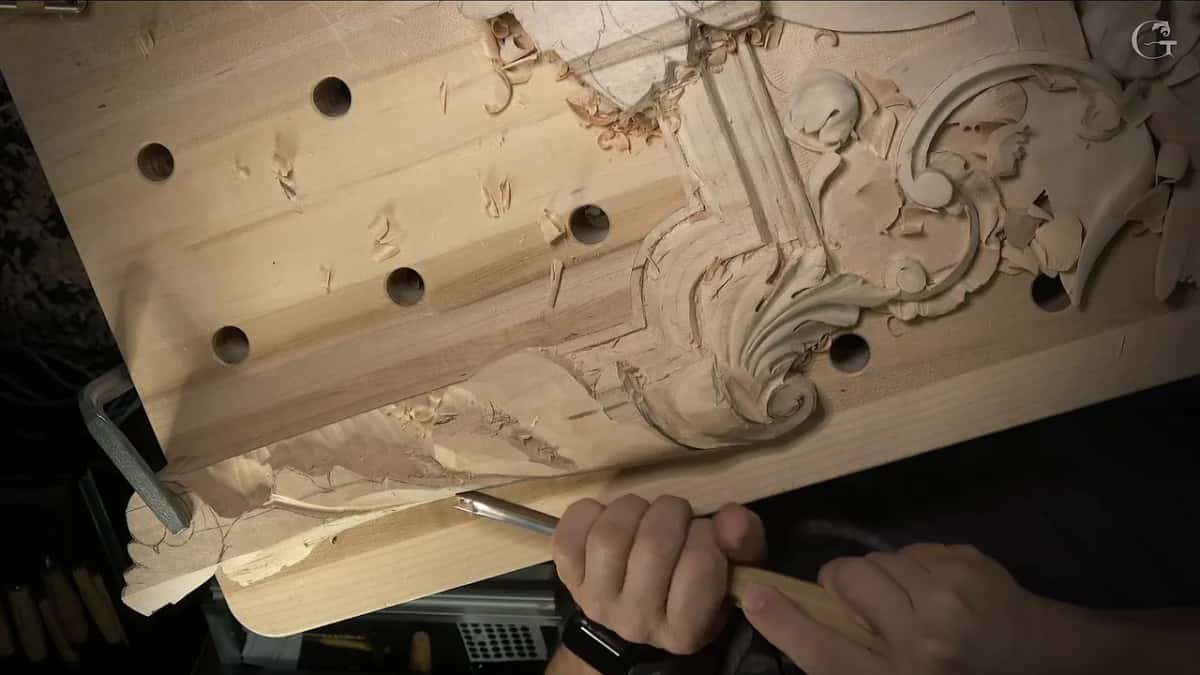 Both hands drive a gouge along the lower band toward the carved leaf, chips near the bench holes