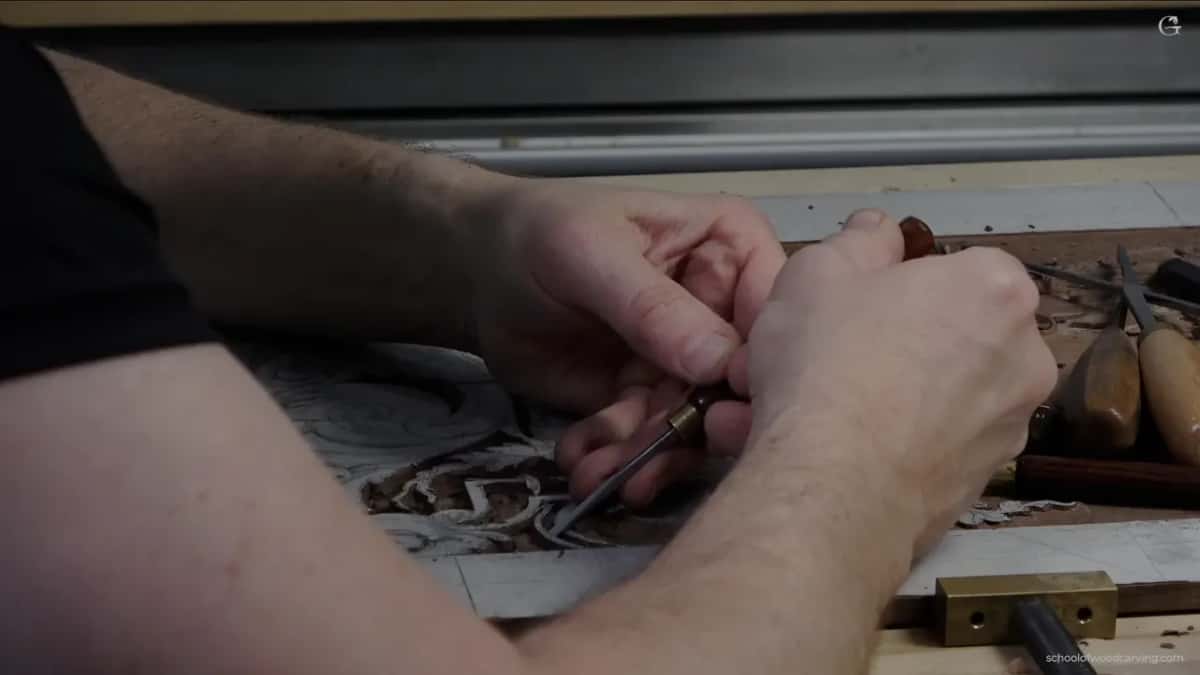 Two hands guide a small gouge across a panel, a brass block and tool handles close by