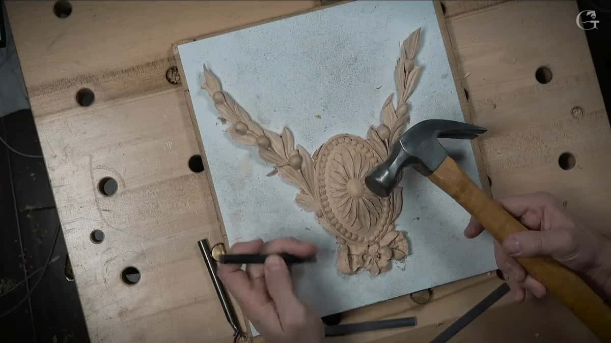 A claw hammer and a punch over a carved oval flower and garland on a white board.