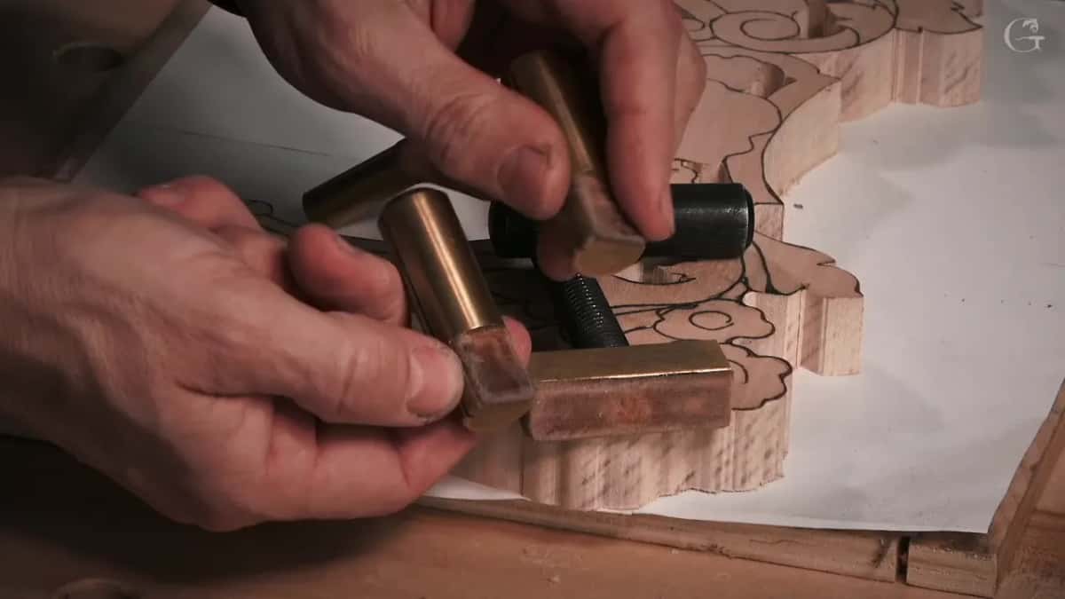 Two hands fit a brass peg against the edge of a roughed carving, a black screw nearby