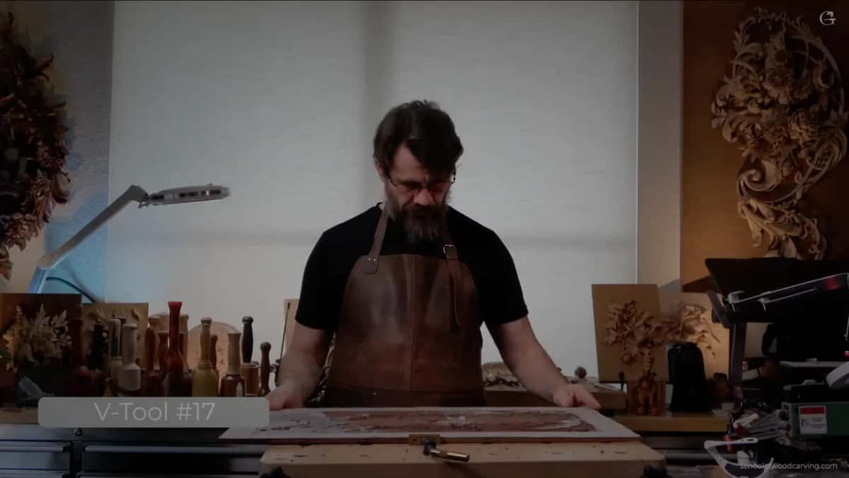 Head down in a brown apron at the bench, hands on a long panel, a caption reading V-Tool #17