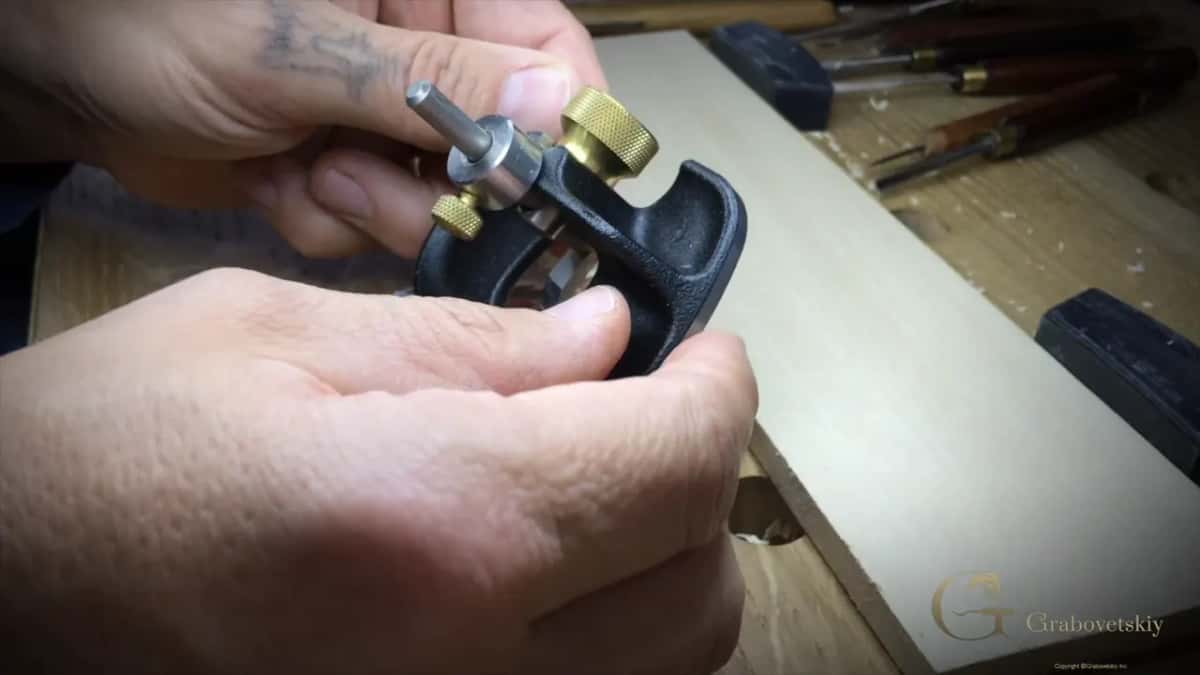 Router plane held up in both hands, two brass knobs on top, carving tools lying behind it on the bench.